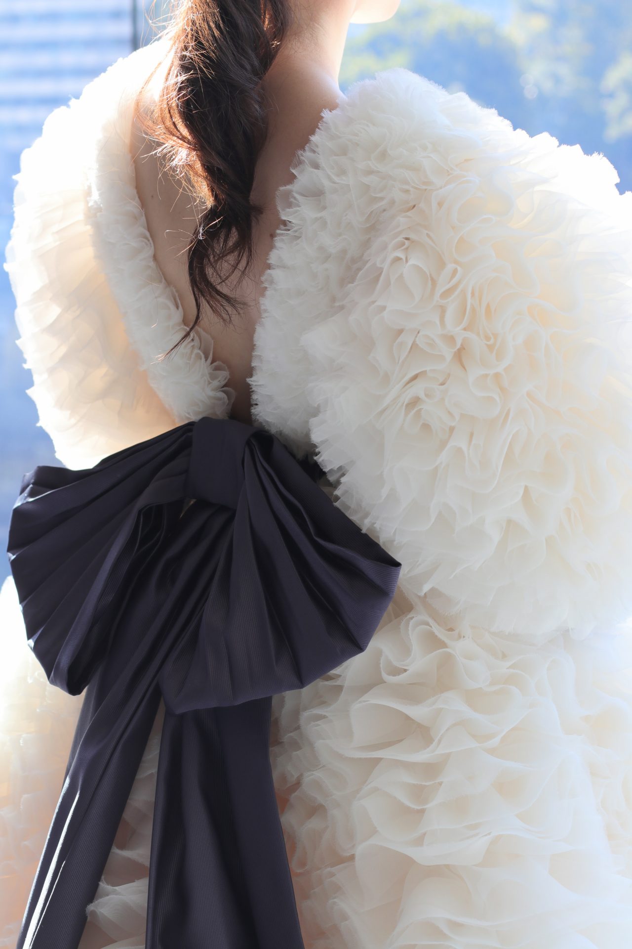 花嫁の美しいバックスタイルを際立たせるVバックのウェディングドレスは、幾重にも重なるふんわりとした繊細なオーガンジー素材のフリルと、ダークパープルの大振りなリボンが女性らしい可愛らしさとエレガントさを引き立てるデザインです