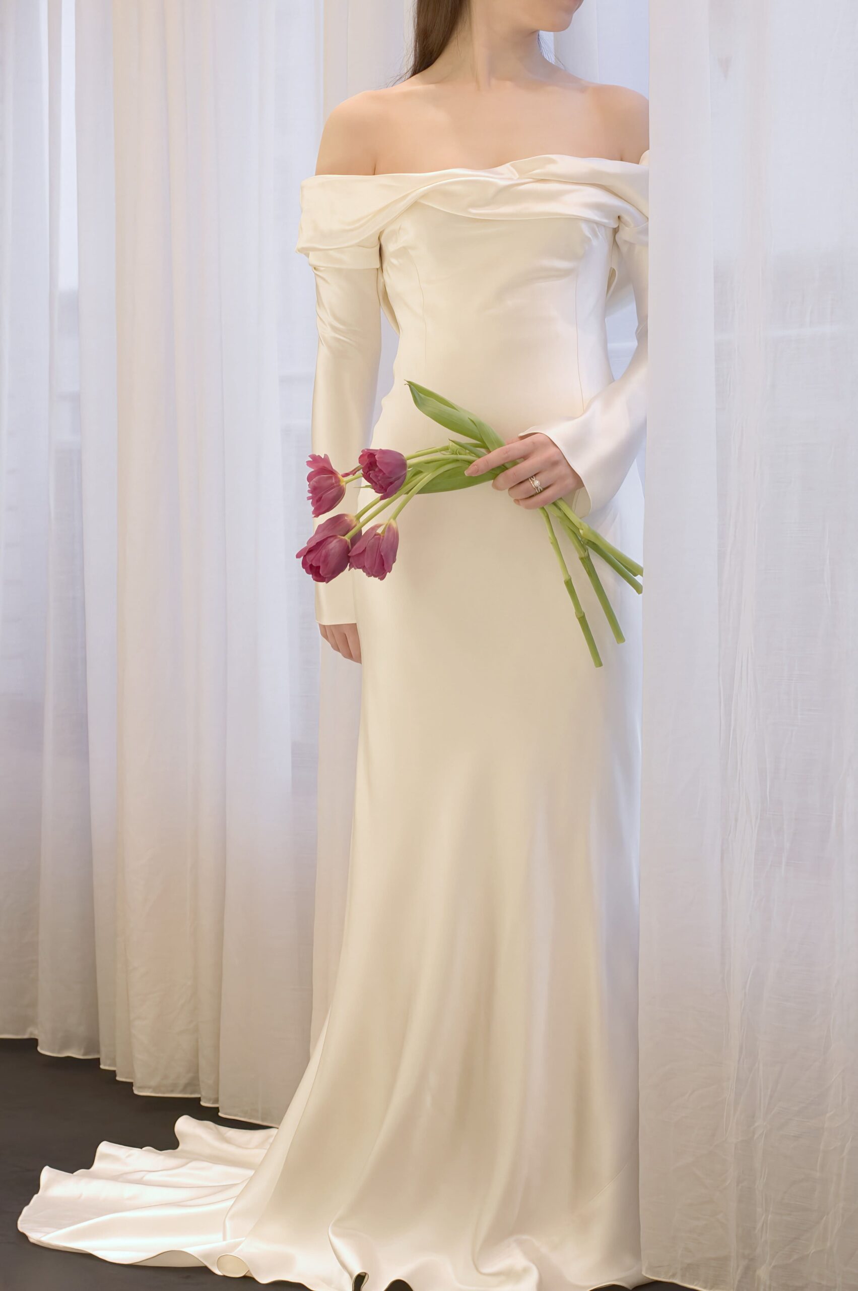 春の結婚式におすすめのウェディングドレスは、シルクの艶めきが美しい細身のシルエットで、オフショルダーがデコルテを華奢にみせるロングスリーブのデザインです