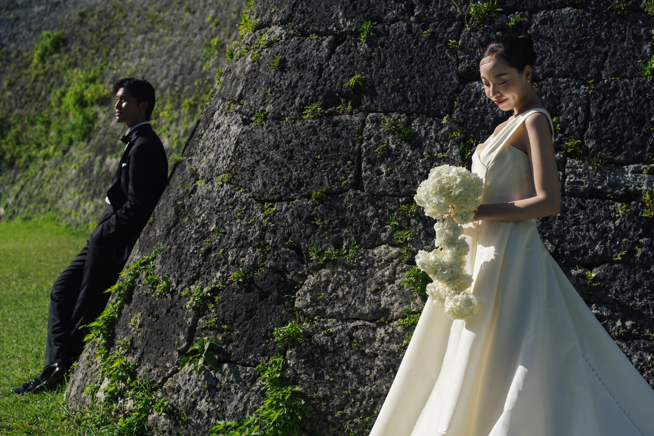 まとめたシニヨンスタイルで白いあじさいの花のブーケをもちアムサーラのウェディングドレスを着用したプレ花嫁とブラックのタキシードを着用した新郎のトリート沖縄店のフォトウェディングでの撮影
